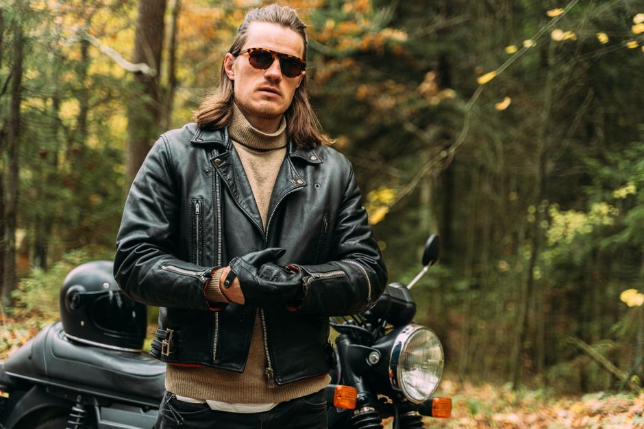 Confident biker in black leather jacket standing by motorcycle in a forest setting.