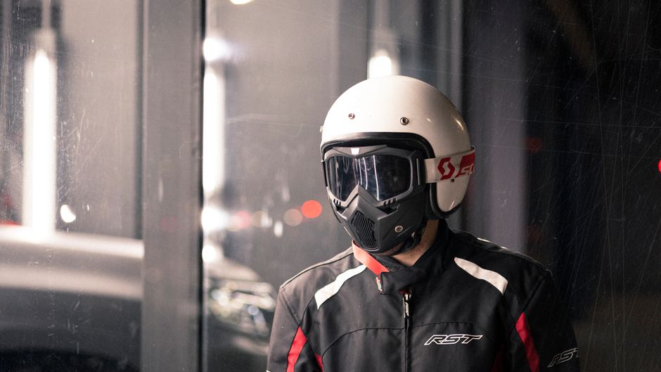 Motorcyclist wearing protective gear stands indoors. Nighttime repair shop setting.